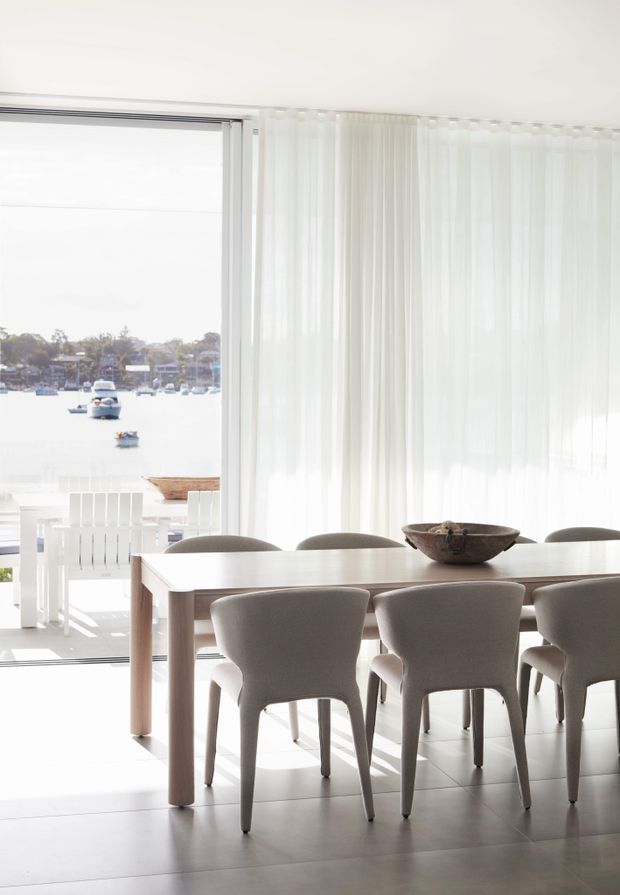 A bright, airy dining room with a long light-wood dining table and upholstered taupe-grey chairs. Floor-to-ceiling sheer curtains frame expansive glazing and an open waterfront view, while white tiled flooring and minimal decor create a clean, refined atmosphere.