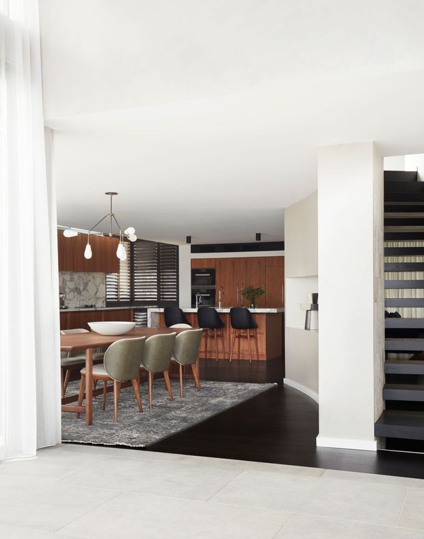 Bright open-plan interior showing a dining area connected to a compact kitchen and bar, with a dark timber floor, warm timber cabinetry, marble-look backsplash, sculptural pendant lighting, and a black open-riser staircase. The foreground has pale tiled flooring and sheer curtains, creating a light contemporary residential setting.