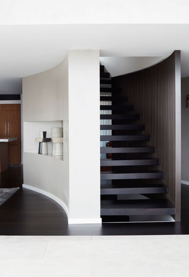 A refined interior foyer centered on a dramatic open-riser staircase with dark timber or metal treads and a curved vertical-slat balustrade. A sweeping curved plaster wall with recessed display shelving divides the stair from a darker adjoining space. The palette is monochromatic, combining white walls and pale flooring with dark timber floors, stair elements, and joinery.