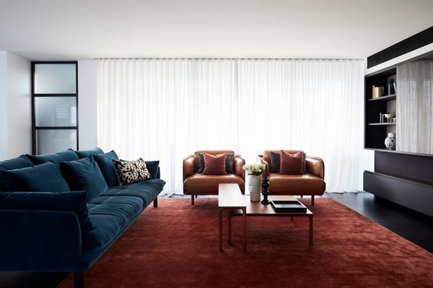 A refined contemporary living room with a deep teal sectional sofa, two cognac leather armchairs, and nested rectangular coffee tables arranged on a large rust-red rug. Full-height sheer curtains provide soft daylight, while dark built-in shelving and cabinetry with timber-toned panels line one wall. Black-framed glazing and a restrained palette create a polished, slightly dramatic interior.