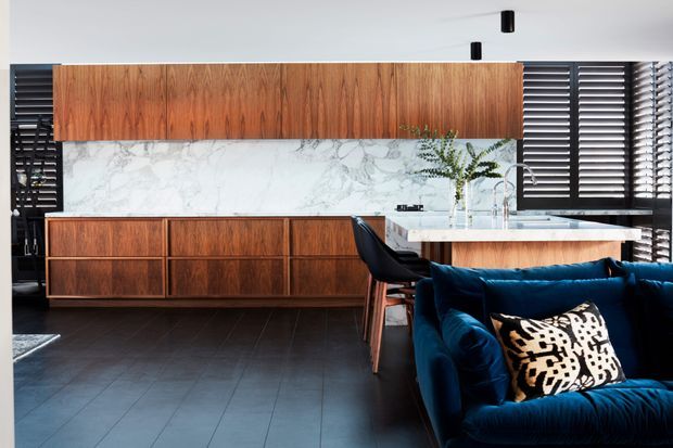 A sophisticated open-plan kitchen and living area featuring extensive warm timber cabinetry, a white marble-look splashback, dark tiled flooring, a white island bench, black bar stools, and a deep navy velvet sofa. Dark plantation shutters and black accents create a strong, refined contrast.