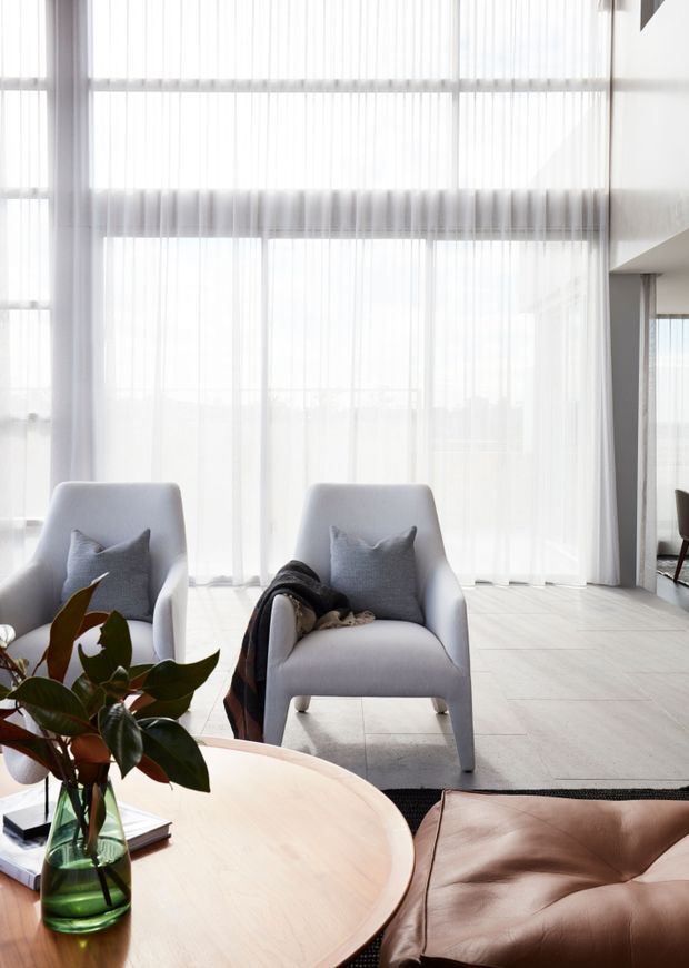 Bright contemporary living room with two light grey upholstered armchairs, a round pale timber coffee table, a brown leather ottoman or sofa seat, and a leafy plant in a glass vase. Floor-to-ceiling sheer curtains span large glazed openings, creating an airy, softly lit interior with pale flooring and minimal architectural detailing.
