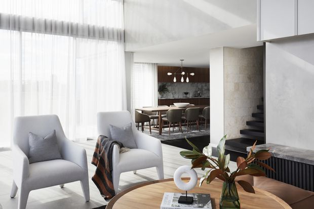 Bright, refined contemporary open-plan living space with two upholstered armchairs in the foreground, a round timber coffee table, and a dining area beyond. Floor-to-ceiling sheer curtains provide abundant natural light, while a dark timber feature wall, stone-clad stair volume, and black stair treads add contrast. A sculptural pendant hangs over the dining table, with greenery and minimal decorative objects providing subtle accents.