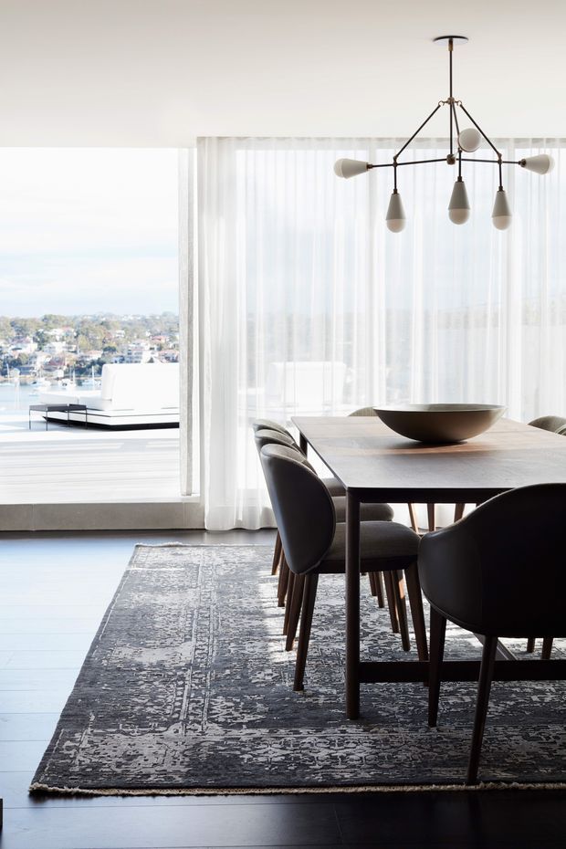 Bright, refined dining area with a dark timber dining table and upholstered dining chairs arranged on a large patterned rug. Sheer white curtains frame expansive glazing, with a sculptural multi-arm pendant light overhead and a large bowl centerpiece on the table. A light-filled living area and distant waterfront suburb are visible beyond the glazing.