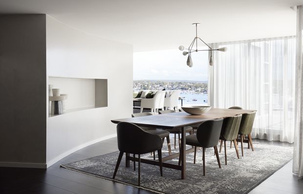 A refined contemporary dining area with a dark timber dining table, sculptural pendant light, upholstered dining chairs, and a large patterned rug. White walls feature a recessed display niche, while sheer floor-to-ceiling curtains open to expansive waterfront views.