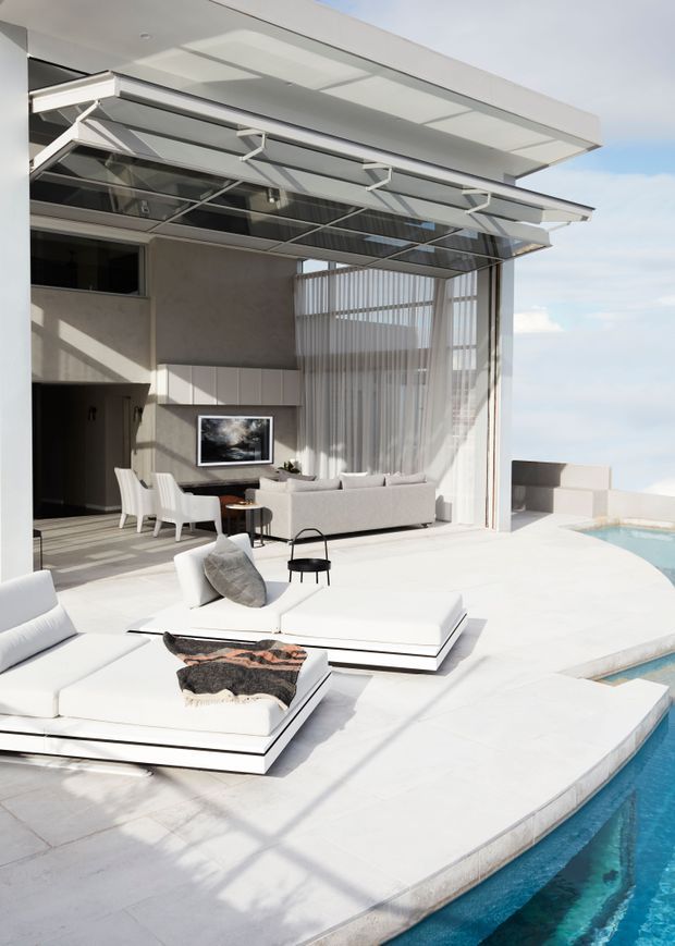 A bright, luxurious modern outdoor pool terrace adjoining an open-plan living and dining area. The space features a white sculptural pool deck, turquoise swimming pool, large white modular sun loungers, a pale sectional sofa, white dining chairs, expansive glazing with sheer curtains, and a large retractable glass-and-metal awning. The architecture has crisp horizontal lines, a flat roof, exposed structural elements, and a seamless indoor-outdoor connection.