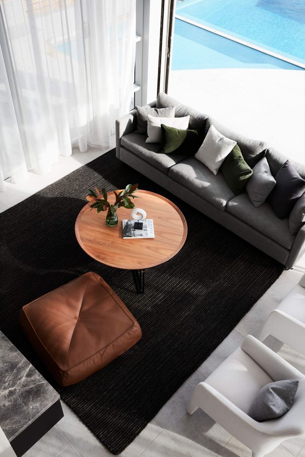 Bright, high-angle view of a refined living room arranged around a round timber coffee table. The room features a large grey sofa with white and olive cushions, a black textured rug, a brown leather ottoman, and a white lounge chair. Sheer curtains and expansive glazing open onto a bright blue swimming pool, while a dark stone-clad fireplace or plinth is visible at the edge of the room.