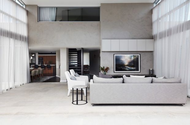 Bright, spacious contemporary living room with a large light-grey sectional sofa, white lounge chairs, and a small round side table arranged around a wall-mounted artwork and low media console. Full-height sheer curtains line both sides of the double-height space, with an upper-level interior window and an open connection to a dining or kitchen area at the rear. Pale textured walls and light flooring create an airy, refined interior.