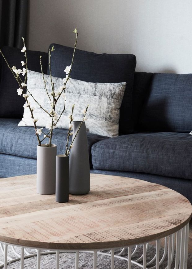 Close-up interior vignette of a contemporary living room featuring a dark blue-grey upholstered sectional sofa with contrasting textured cushions, three minimalist cylindrical vases holding delicate branches, and a large light timber round coffee table with a white curved metal base. Neutral grey walls and a textured rug complete the restrained, modern setting.