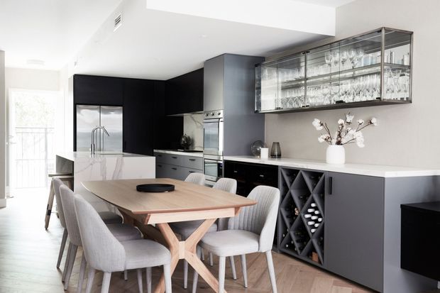 Contemporary open-plan kitchen and dining area with a large white island, dark matte cabinetry, integrated stainless-steel appliances, a pale engineered-stone benchtop, and a timber dining table surrounded by upholstered dining chairs. A glass-fronted display cabinet, wine storage, and barware shelving form a feature wall, while bright glazing brings in natural light.
