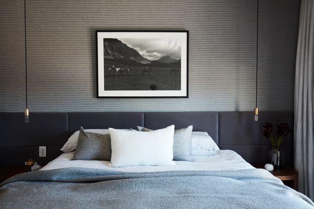 A contemporary bedroom centered on a neatly made bed with layered white and grey bedding, a dark upholstered full-width headboard, textured grey wall covering, and a framed black-and-white mountain photograph. Slim pendant lights hang on either side of the bed, with dark bedside tables, a small vase, and full-height curtains completing the calm, refined scheme.