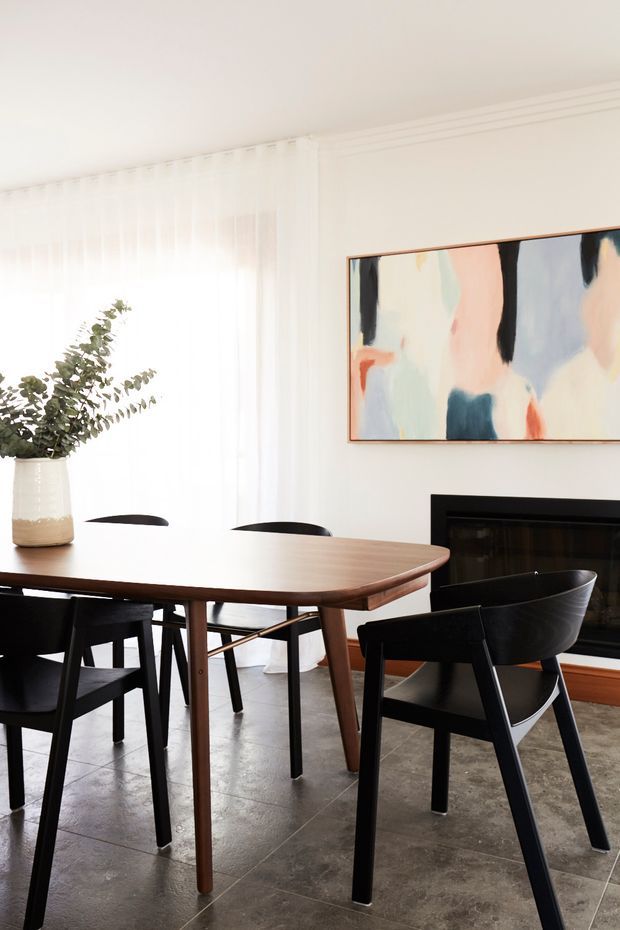 Bright, refined dining room with a large rounded-edge timber dining table and black dining chairs, positioned in front of a dark fireplace. Sheer white curtains provide abundant natural light, while a large abstract artwork and simple vase arrangement add visual interest. The space has a clean contemporary feel with warm timber accents and a polished concrete-look tiled floor.