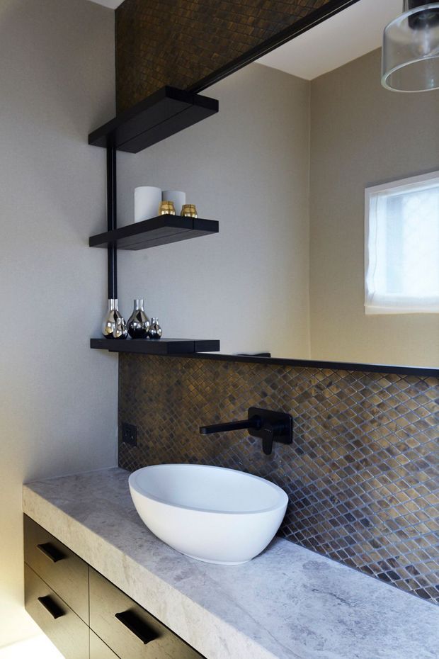 Contemporary bathroom vanity with a white vessel basin set on a pale stone benchtop, dark built-in cabinetry, matte black wall-mounted tapware, open black display shelves, and a striking bronze-brown mosaic tile splashback. A small frosted window provides natural light.