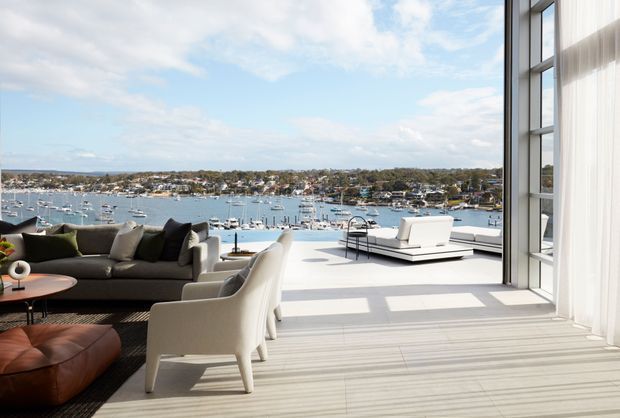 A bright, expansive contemporary living room opening onto a waterfront terrace, with floor-to-ceiling glazing framing a marina and coastal residential outlook. The room features a large charcoal sectional sofa, pale upholstered lounge chairs, a low round timber coffee table, and light timber flooring. Strong daylight creates a calm, airy, luxurious atmosphere.