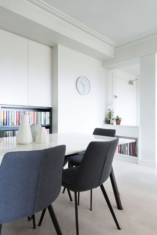 Bright, refined dining room with a white rectangular dining table, dark upholstered dining chairs, built-in white cabinetry and open bookshelves, decorative objects, and a minimalist wall clock. The room features crisp white walls, ornate ceiling cornices, recessed architectural detailing, and pale carpeting.