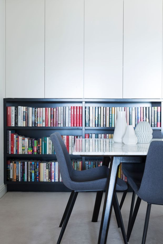 A bright, modern study or dining workspace with a full-height dark built-in bookshelf filled with books, a pale vertically panelled wall above, and a light rectangular table surrounded by dark upholstered chairs. White sculptural vases sit on the tabletop, creating a clean, refined contemporary interior.