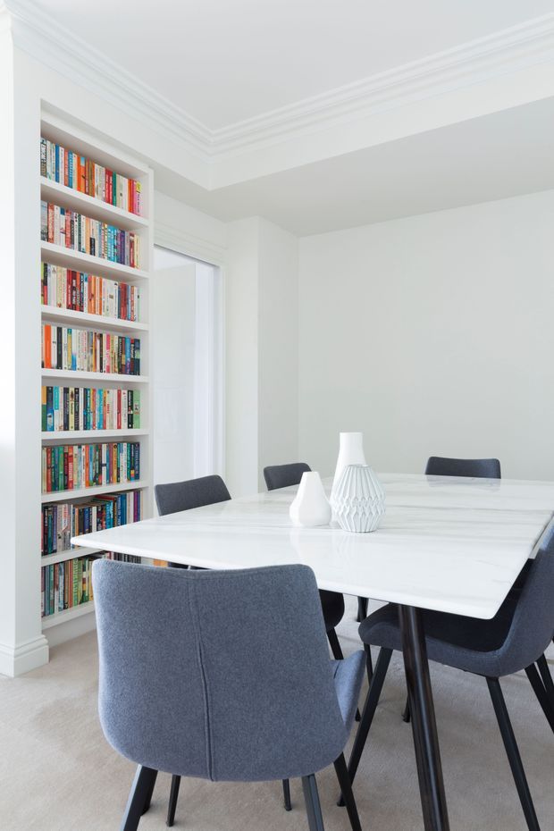 Bright, refined dining room with a large white marble-look rectangular dining table, dark grey upholstered dining chairs, white sculptural vases, and a full-height built-in bookshelf filled with books. The room has white walls, ornate crown moulding, light carpet, and generous natural light.