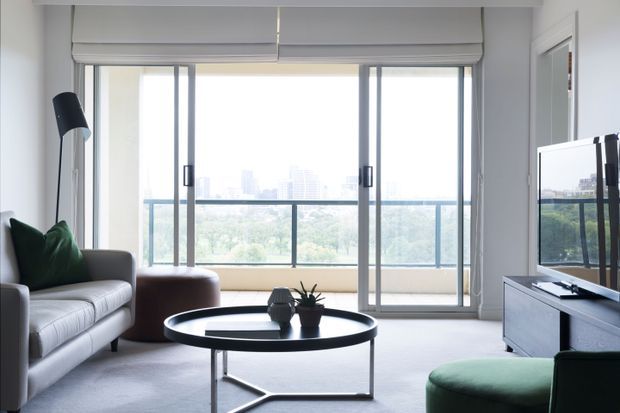Bright contemporary living room with a large sliding glass opening onto a balcony and distant city view. The room features a grey sofa, dark green accent cushions and ottoman, a low black round coffee table, a floor lamp, and a wall-mounted television with a low media console. Roller blinds and slim grey window frames reinforce the clean modern look.