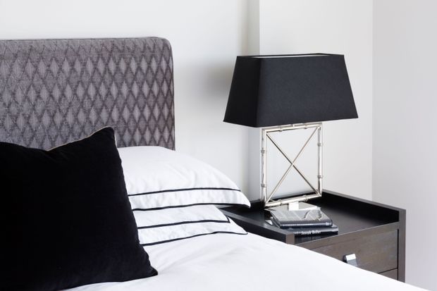 A close-up of a refined bedroom corner featuring a bed with a grey diamond-patterned upholstered headboard, crisp white bedding with black piping, a black accent cushion, and a dark bedside table. A black-shaded lamp with a metallic crossed-frame base sits on the table against white walls.