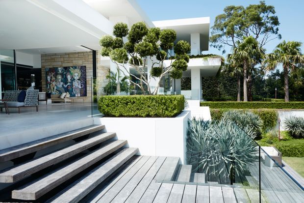 Bright contemporary residential exterior showing a white flat-roofed house with extensive open glazing, a covered outdoor entertaining area, broad timber decking and steps, manicured hedges, sculpted topiary, and drought-tolerant landscaping. The view looks toward the house from the landscaped garden and deck.