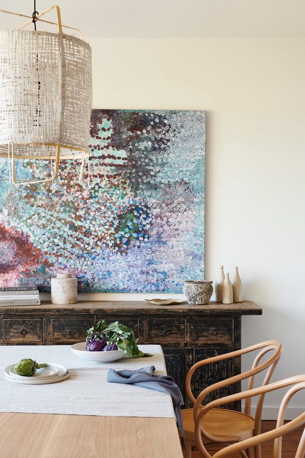 Warm, refined dining area featuring a large light timber dining table with a white tablecloth, curved timber dining chairs, and a dark rustic timber sideboard. A large abstract artwork in blue, mauve, grey, and burgundy tones forms the main backdrop, complemented by a woven pendant light, ceramic vessels, greenery, and simple tableware.
