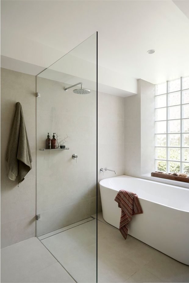 Bright, minimalist bathroom with a frameless glass walk-in shower, large pale stone-look wall and floor tiles, freestanding white bathtub, chrome shower fittings, and a large glass-block window providing diffused natural light. Neutral finishes and simple fixtures create a clean, calm atmosphere.