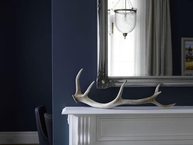 Close-up of a refined fireplace mantel with white painted classical moulding and fluted detailing against a deep navy wall. A natural antler sculpture rests on the mantel, while an ornate dark-framed mirror reflects a bright window and hanging light. A portion of a dark upholstered chair is visible at left.