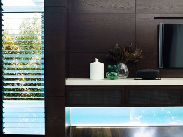 Close-up interior view of a contemporary living room media wall, featuring dark timber wall panelling and cabinetry, a wall-mounted television, a light stone or solid-surface shelf, decorative objects, and a bright glazed or illuminated lower feature panel. Horizontal louvred blinds on the left admit daylight and reveal greenery outside.