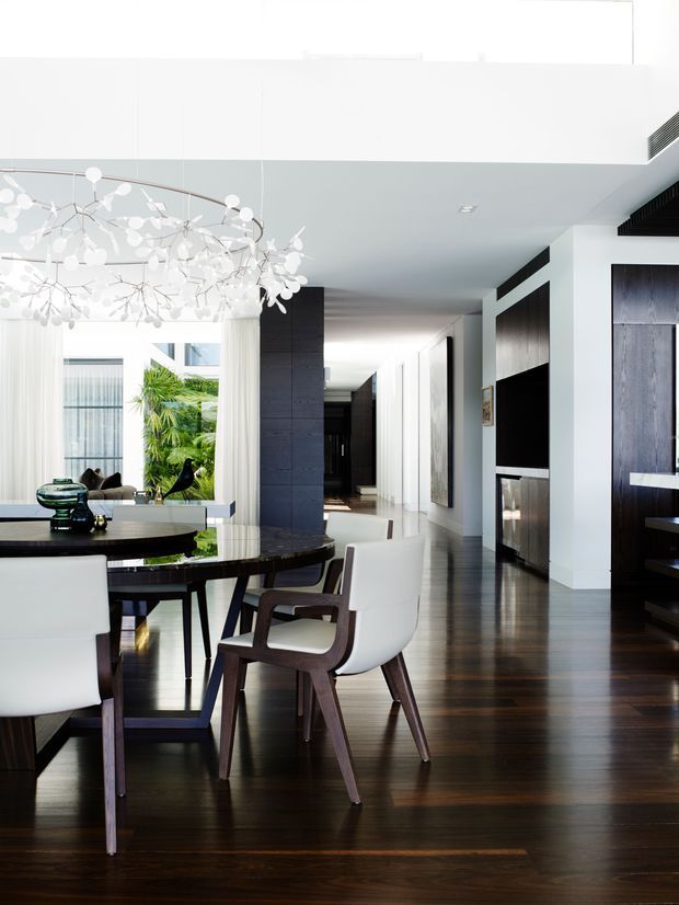 Bright, high-end open-plan interior featuring a formal dining area with a dark oval table, white upholstered and timber dining chairs, and a sculptural branching pendant light. The space extends into a sleek hallway and living area with white walls, dark timber flooring, dark feature joinery, large windows, and tropical greenery visible outside. The overall design is clean-lined, refined, and contemporary.