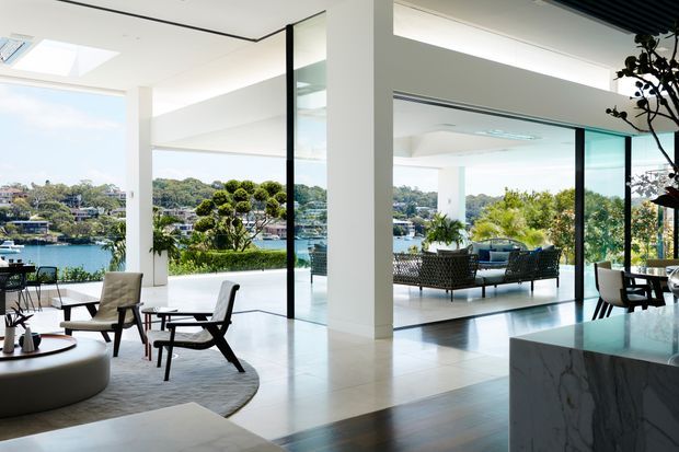 Bright contemporary open-plan interior overlooking a leafy waterfront setting. The space features floor-to-ceiling glazing, white structural columns and walls, pale tiled flooring, dark timber flooring, a marble-clad kitchen island, and an adjoining living area with modern lounge furniture. A large covered terrace and dining area are visible through the glazing.
