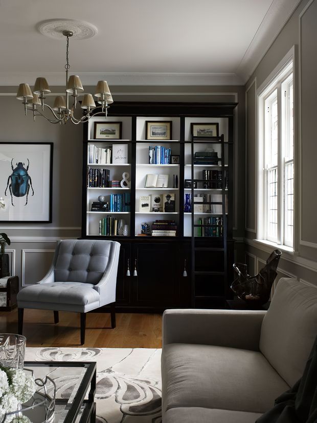 A refined traditional living room with dark grey paneled walls, white trim, built-in black shelving and cabinetry filled with books and framed art. The room features a tufted grey armchair, a large upholstered sofa, a sculptural chandelier, hardwood flooring, a patterned area rug, and tall multi-pane windows providing natural light.