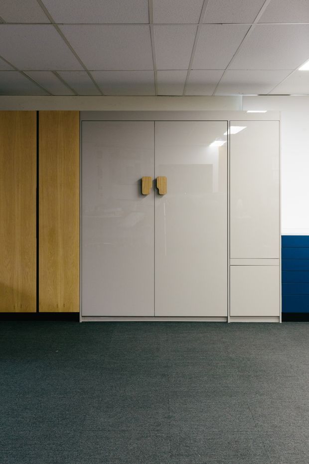 A minimalist commercial or office interior showing a wall of built-in storage cabinetry. The cabinetry combines tall natural timber-look panels, glossy off-white doors with light wood handles, and a partial blue drawer unit. The room has dark grey carpet flooring and a suspended acoustic ceiling with recessed fluorescent lighting.