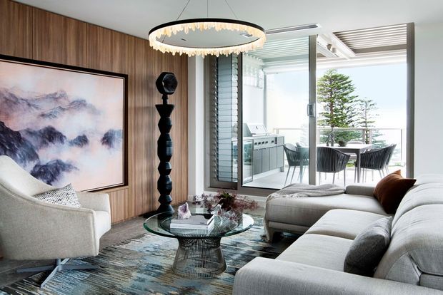 Contemporary living room with a large light-grey sectional sofa, sculptural lounge chair, round glass coffee table, textured area rug, timber wall panelling, oversized artwork and a statement pendant light. Full-height glazing opens the room visually to an outdoor dining terrace and compact outdoor kitchen, with a coastal view beyond.