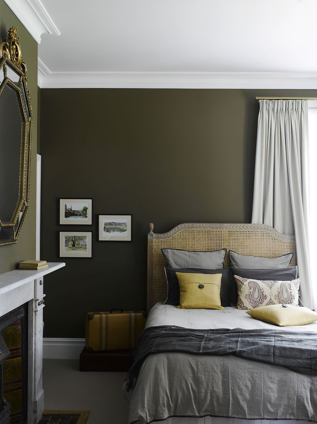 A refined bedroom with dark olive-grey walls, crisp white ceiling cornices and skirting, and a traditional white mantelpiece. The bed features a woven cane headboard, layered grey and patterned cushions, a mustard accent cushion, and a charcoal throw. An ornate gilded mirror, framed artwork, white curtains, and a vintage suitcase add an eclectic, classic character.