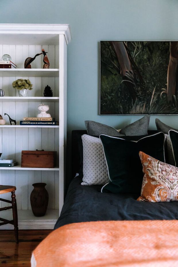 A warmly styled bedroom with a dark upholstered bed, layered decorative pillows, and a burnt-orange throw. A white built-in display bookshelf with vertical panel backing occupies the left side, while a blue-grey wall and framed nature artwork provide the backdrop. Timber flooring, traditional furniture details, and eclectic decorative objects create a refined, cozy atmosphere.