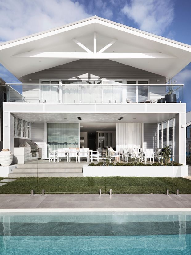 Front exterior view of a bright two-storey contemporary residence with a broad white gabled roof, grey horizontal cladding, expansive glazed openings, upper balcony, covered outdoor dining area, landscaped lawn, and swimming pool in the foreground. The facade includes strong geometric triangular roof detailing and white-framed glazing.