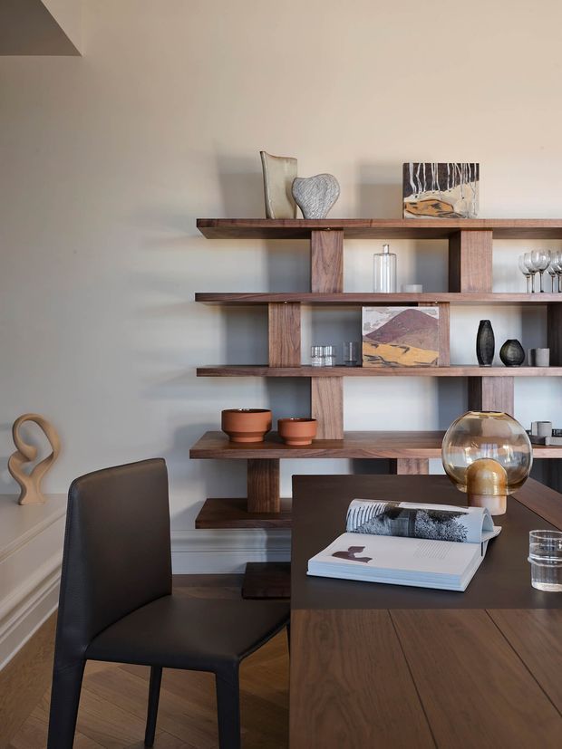 A refined dining and display area with a dark timber dining table and upholstered dining chair in the foreground. An open, built-in timber shelving unit spans the rear wall, displaying ceramics, glassware, artwork, and sculptural objects. The room has warm wood flooring, soft neutral walls, restrained decorative styling, and a contemporary yet mid-century-influenced character.