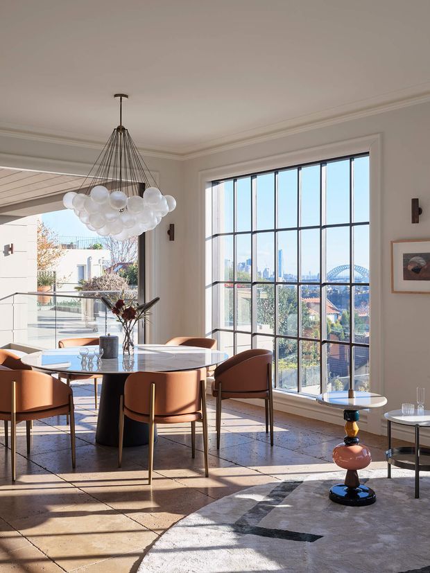 Bright, refined dining area with a round dining table and sculptural upholstered chairs, set beside expansive black-framed grid windows. A dramatic clustered globe chandelier hangs above the table, while a large rug, artwork, side table and modern decorative lighting add layered detail. The windows overlook a city skyline and a distinctive arched bridge, with an adjacent outdoor terrace visible through the opening.