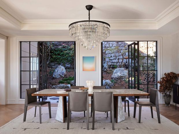 Elegant dining room with a large rectangular stone-topped dining table, upholstered dining chairs, a crystal chandelier, pale walls with decorative crown moulding, and wide black-framed glazed doors opening to a rocky landscaped courtyard. The room has a refined contemporary-classic feel with warm timber flooring and a large neutral rug.