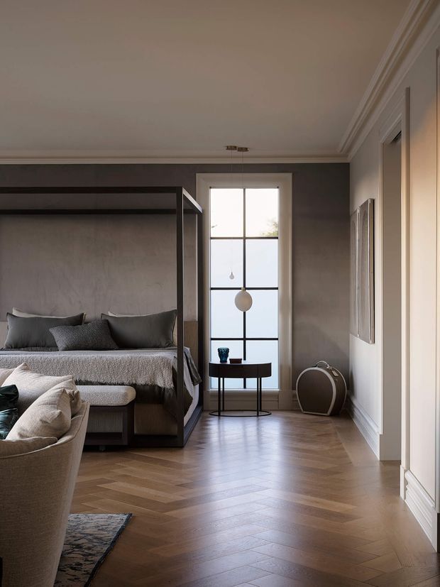 A refined bedroom with a dark-framed four-poster bed, neutral upholstered bedding, and a curved lounge chair in the foreground. The room features warm herringbone timber flooring, taupe-grey walls, white decorative ceiling cornices and skirting, tall divided-pane glazing, and a small round side table with pendant lighting. The overall atmosphere is calm, warm, and sophisticated.