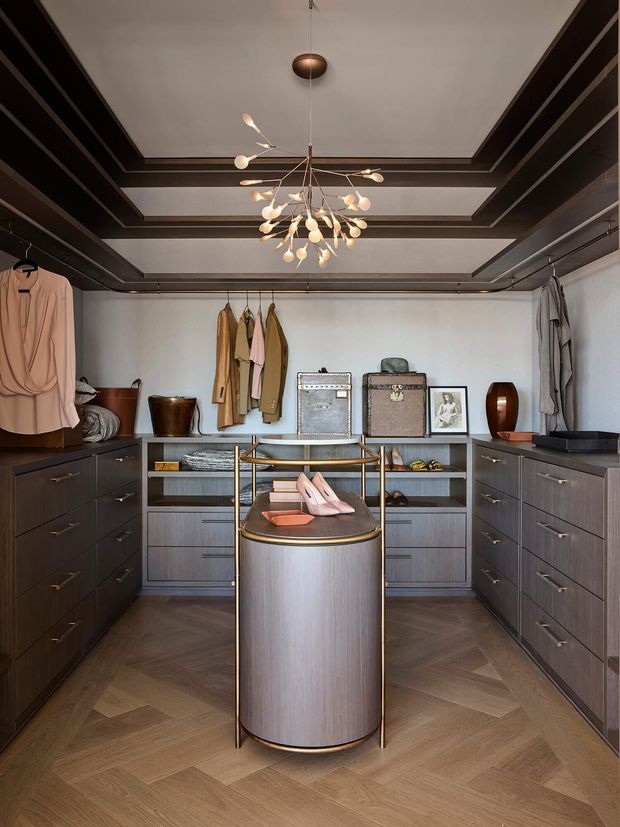 A refined walk-in wardrobe with symmetrical dark wood cabinetry, open shelving, hanging clothes, and a central cylindrical island with brass trim. The room features a warm herringbone timber floor, a coffered ceiling with dark trim, and a sculptural multi-arm pendant light. The overall scheme is dark, warm, and luxurious with carefully styled accessories.