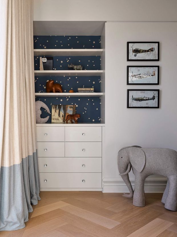 A children's bedroom corner featuring a built-in white dresser and open shelving with a dark navy star-patterned backing, displaying toys and books. Three framed airplane illustrations hang on the pale wall beside it. Full-height two-tone curtains frame the space, while a light timber herringbone floor and a sculptural grey elephant chair add warmth and character.