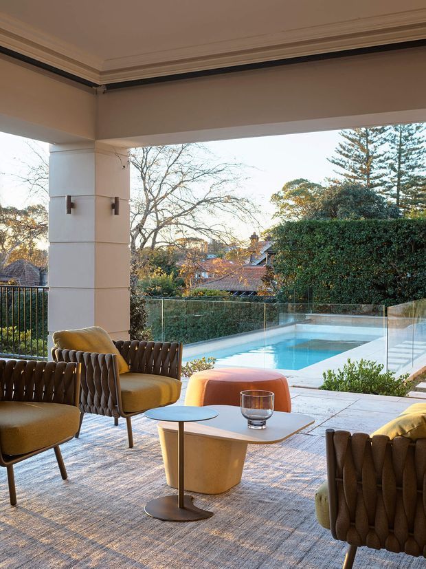 A refined covered outdoor lounge overlooking a rectangular swimming pool and landscaped suburban garden. The space features a broad roof overhang with layered decorative ceiling mouldings, a substantial square column, glass pool balustrading, patterned outdoor carpeting, sculptural upholstered lounge chairs, and a small rounded coffee table. Warm neutral upholstery and timber-toned finishes contrast with the blue pool and greenery beyond.