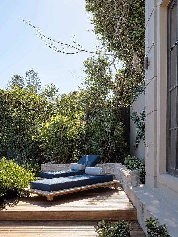 A sunlit landscaped outdoor deck beside a light-coloured rendered building, furnished with a low timber-framed daybed or outdoor lounge in dark navy upholstery. Dense tropical and subtropical planting surrounds a raised timber deck, with a pale stone retaining wall and steps along the garden edge. The scene has a refined coastal-resort feel with strong indoor-outdoor connection.