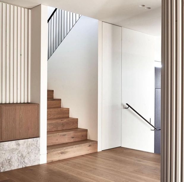 A bright, minimalist residential hallway and stairway with warm timber flooring and open-riser-style timber stairs. White walls and concealed cabinetry create a clean backdrop, while slim black metal stair balustrades and a handrail provide contrast. Vertical timber slats and a textured stone-faced built-in cabinet add architectural detail.