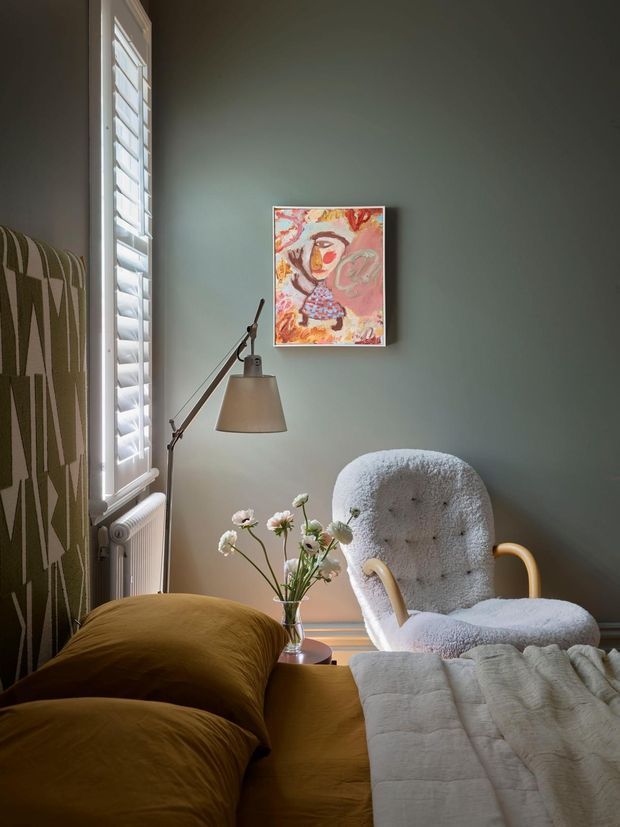 A cozy bedroom corner with a mustard upholstered bed in the foreground, a textured boucle armchair, and a slim adjustable floor lamp. The room has muted deep green walls, a tall window with white plantation shutters, a white radiator, patterned upholstered wall or headboard fabric, and a colourful framed artwork. Soft daylight and layered warm textiles create an intimate, eclectic atmosphere.