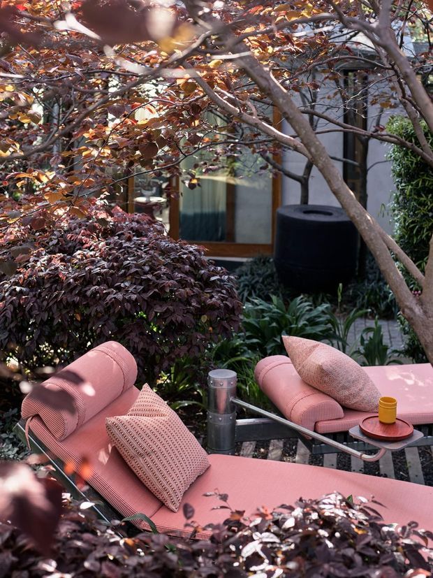 A landscaped outdoor lounge courtyard featuring two pink upholstered chaise lounges with textured cushions, a small round side table, and a dark cylindrical planter. Dense layered planting and mature trees partially screen a modern glazed building facade in the background, creating a secluded garden setting.