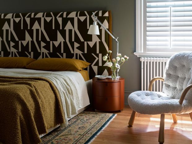 A warmly styled bedroom with a bold geometric black-and-cream upholstered headboard, mustard bedding, textured neutral throw, timber flooring, and a patterned runner rug. A compact reddish-brown bedside table with flowers sits beside the bed, while a sculptural upholstered armchair occupies the sunlit corner near white plantation shutters and a traditional radiator. A slender adjustable floor lamp adds a functional accent.