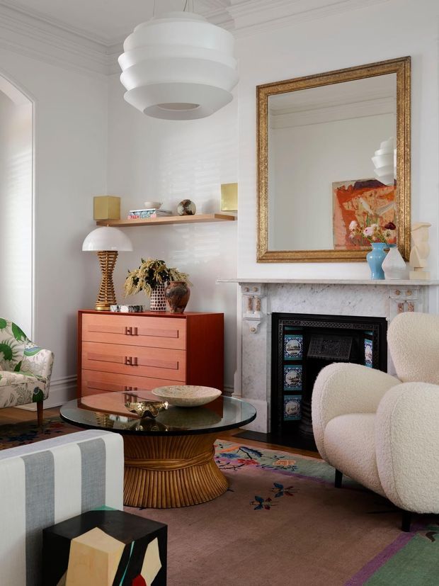A refined living room with ornate traditional architectural details, including decorative ceiling cornices and a carved marble fireplace surround. The room combines a large gilded mirror, mid-century-inspired timber dresser, sculptural pendant and table lamps, a glass coffee table with a fluted wood pedestal, upholstered lounge chairs, and a colourful patterned rug. The palette is predominantly white, cream, warm timber, and muted multicolour accents.
