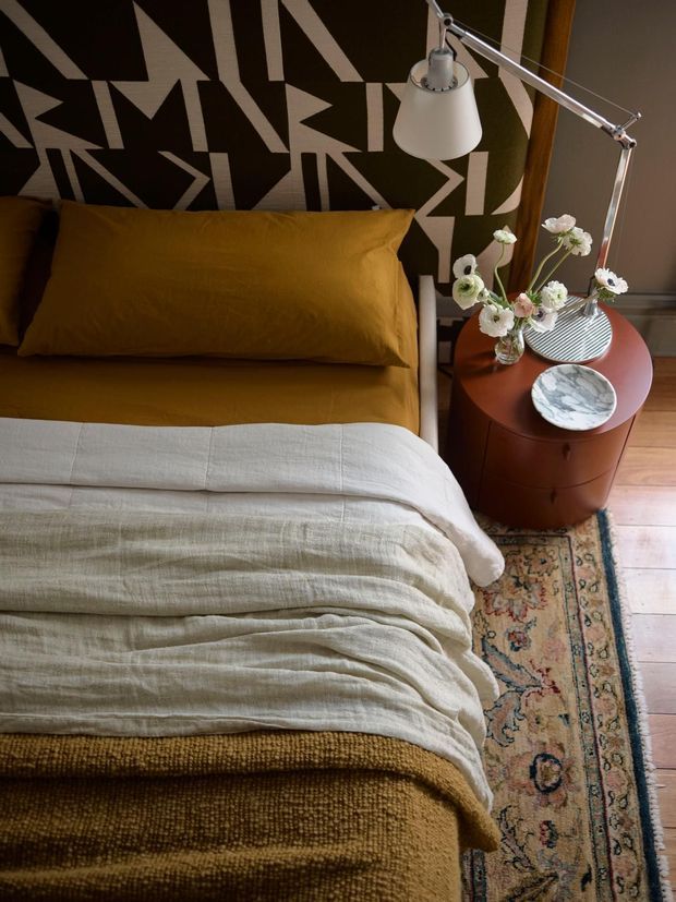 A warmly styled bedroom corner with a mustard upholstered bed and matching pillow, layered off-white bedding, and a bold geometric black-and-cream feature wall. A rounded rust-red bedside table holds a sculptural lamp, vase of flowers, striped dish, and marble bowl. Timber flooring and a patterned vintage-style rug complete the rich, eclectic interior.